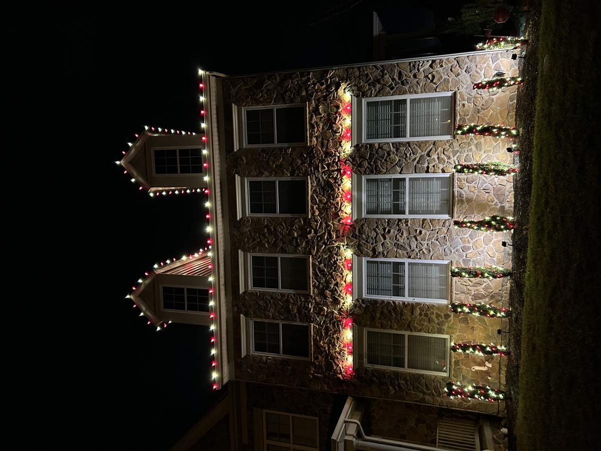 A stone house with an alternating red and warm white roofline and lit garland across the window ledges
